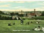Old colour postcard entitled general view of Kidderminster, people in old fashioned clothes sit on the grassed hill with industrial buildings in distance. 