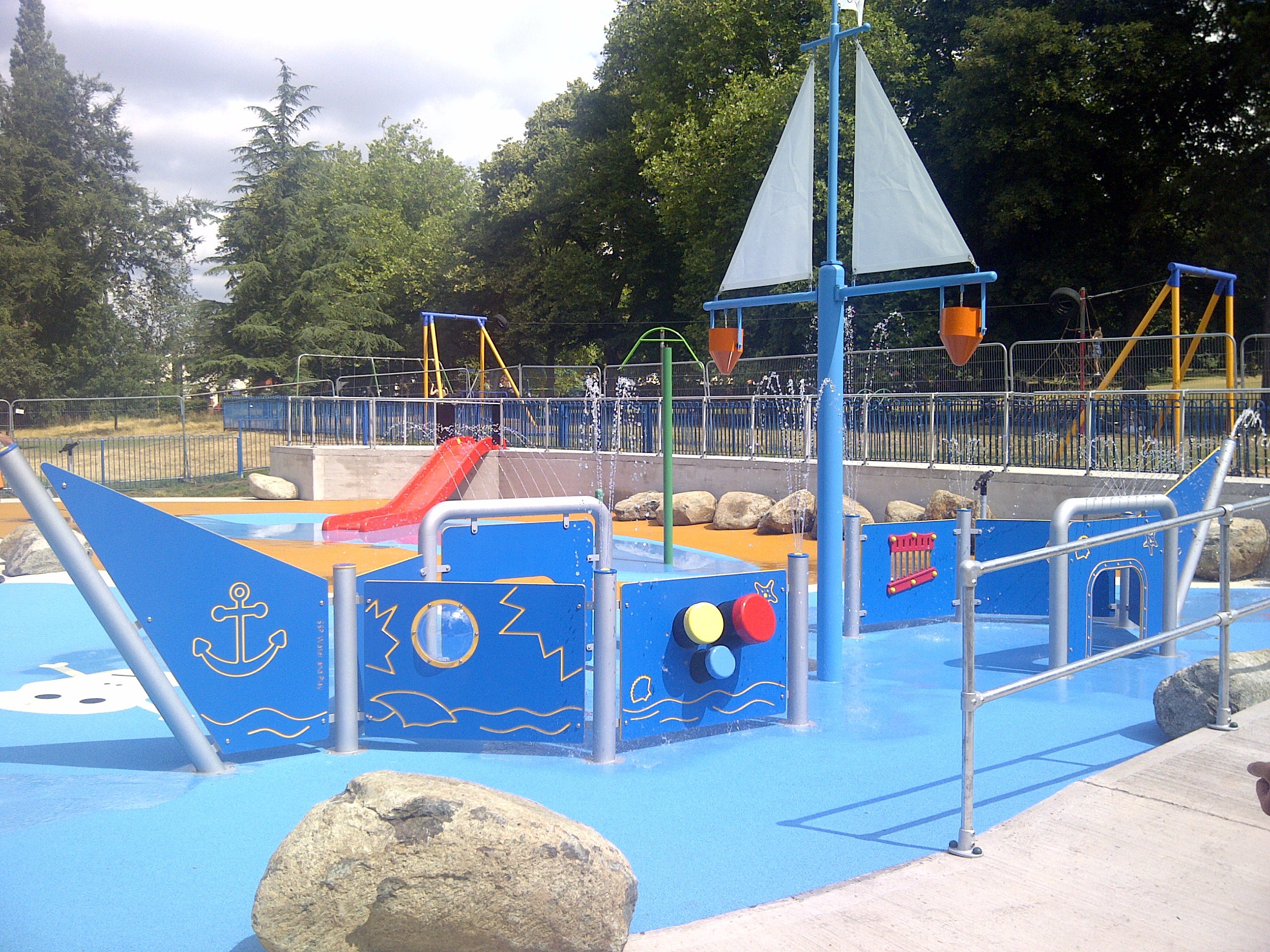 blue floored waterplay area with various sprinklers and water jets some forming a pirate ship shape