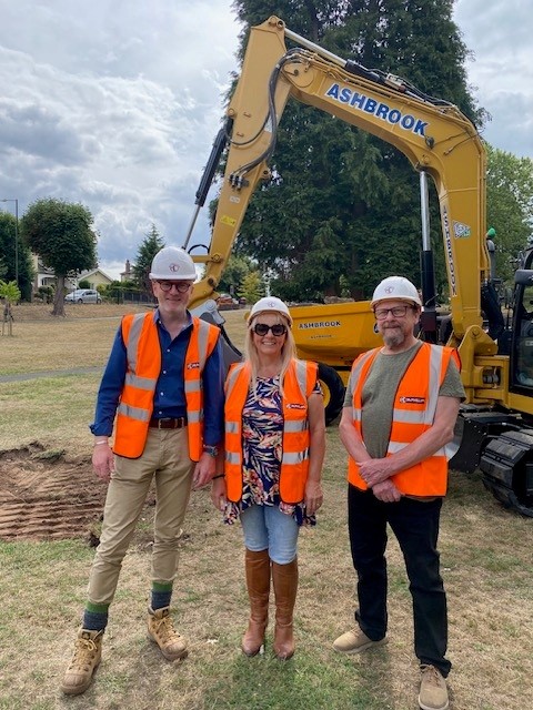 3 people in bright colourted hi-vis vest wearing hard hats with mCHINE DIGGER BEHIND THEM IN LOCAL PARK